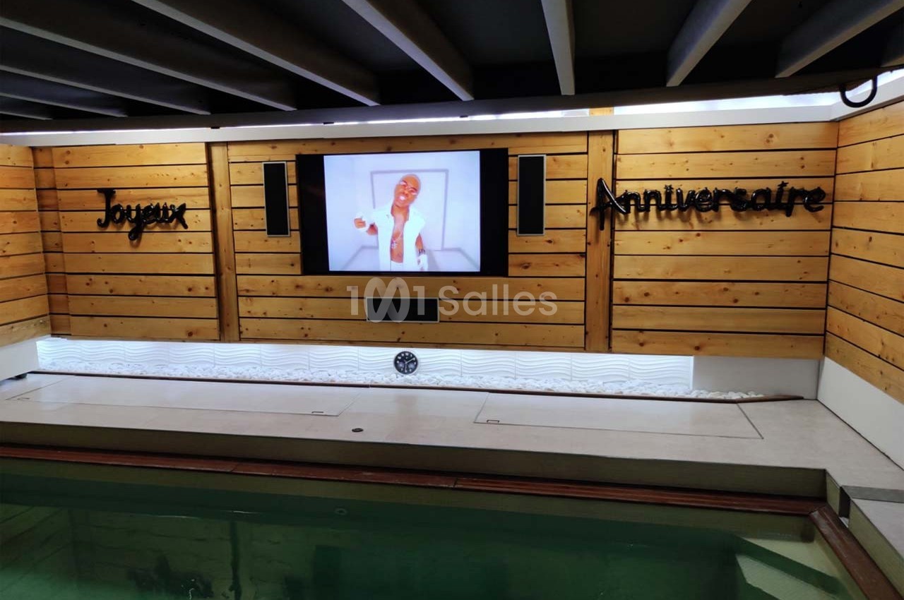 Mur en bois avec un écran affichant une personne souriante, entouré des mots ’Joyeux Anniversaire’, au-dessus d'une piscine.