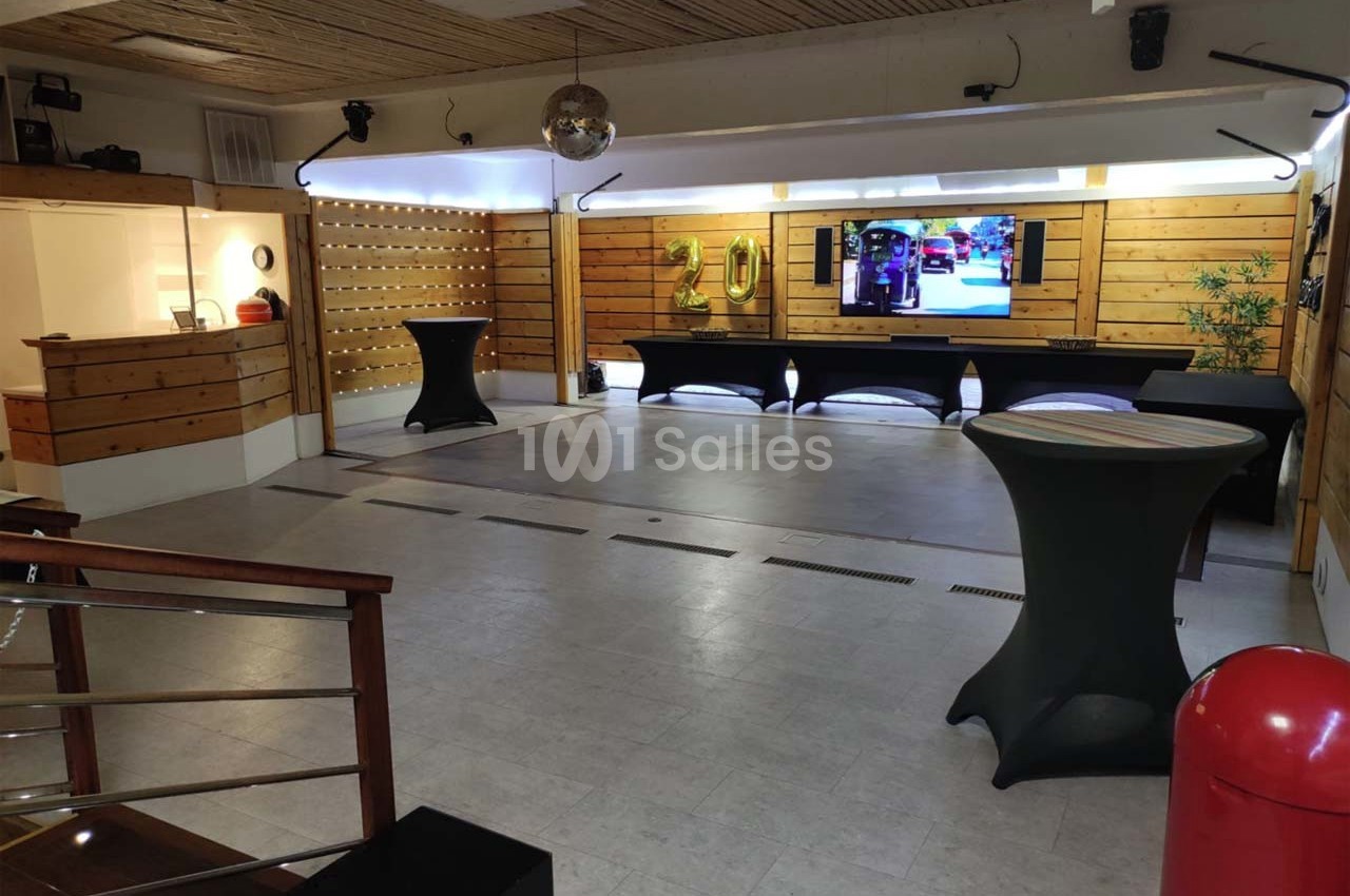 Salle vide avec parquet au plafond, tables hautes habillées de housses noires, écran mural et décoration en bois.