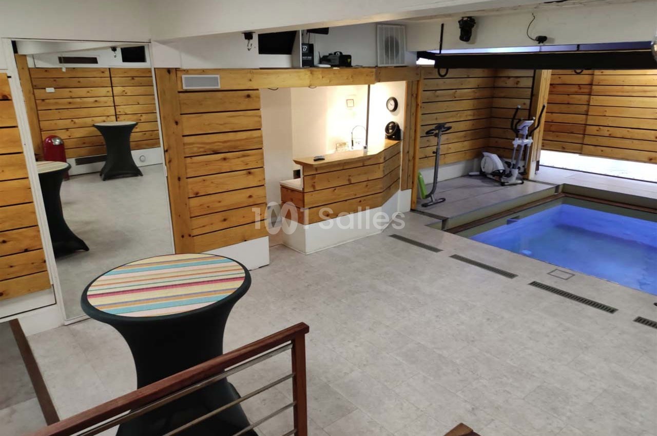 Salle intérieure avec piscine, murs en bois, tables hautes et équipement de sport dans un espace lumineux.