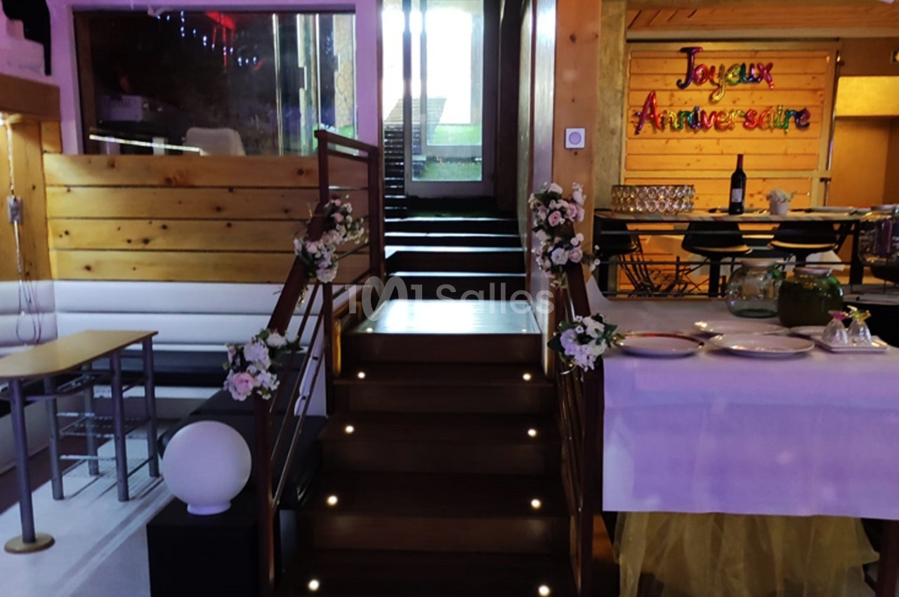 Escalier décoré de fleurs menant à une salle festive avec une table dressée et une bannière ’Joyeux Anniversaire’.