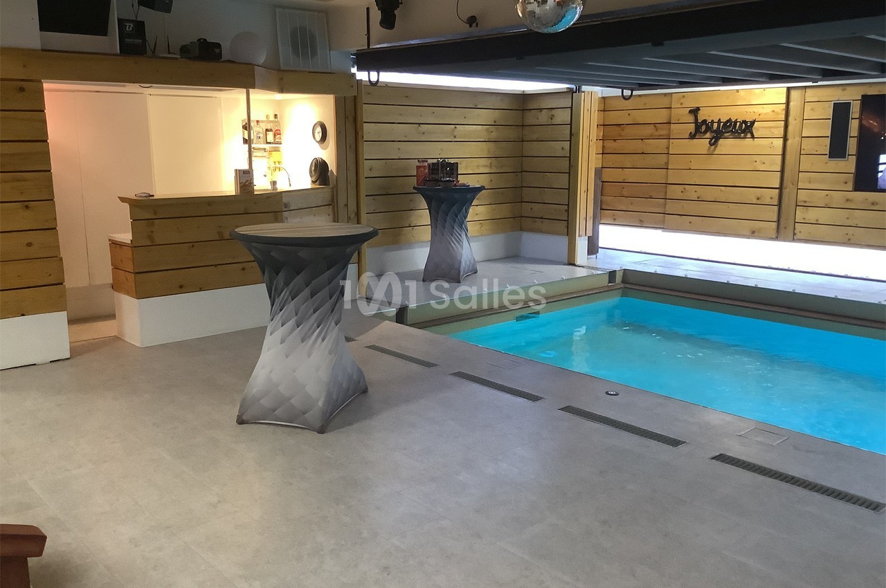 Espace intérieur avec piscine, murs en bois, tables hautes grises et un comptoir éclairé en arrière-plan.
