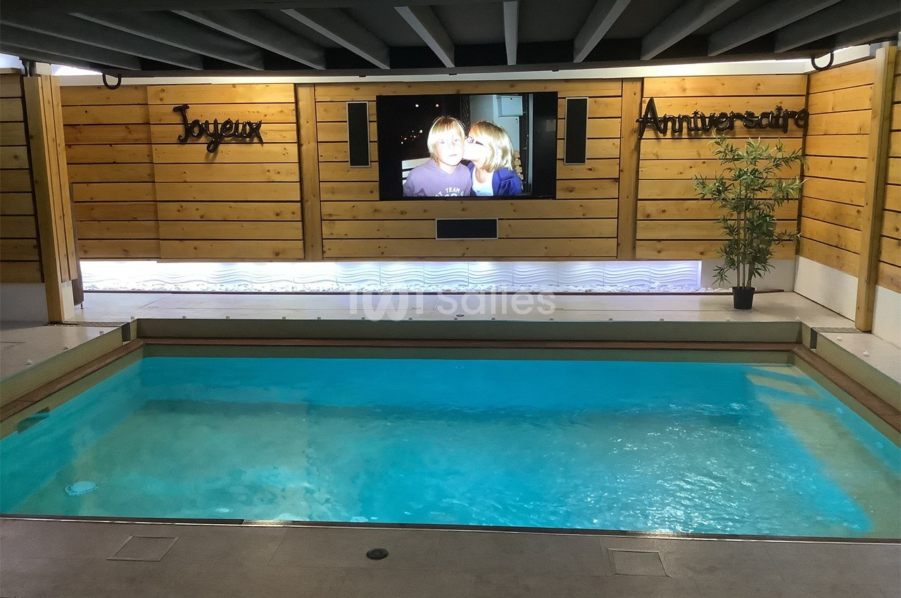 Piscine intérieure éclairée avec un écran mural affichant deux personnes et des panneaux en bois portant l'inscription…