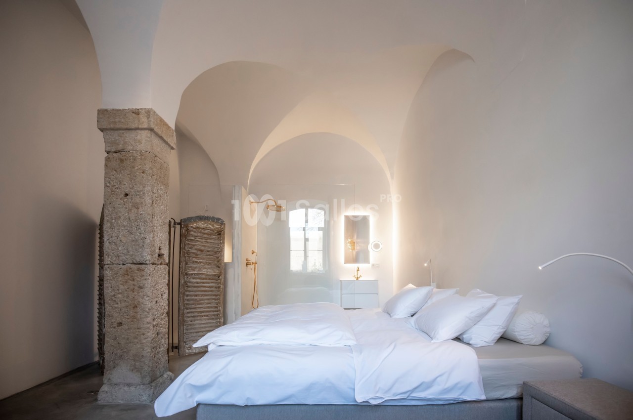 Chambre lumineuse avec un lit double, des murs voûtés blancs, une colonne en pierre et un éclairage moderne.