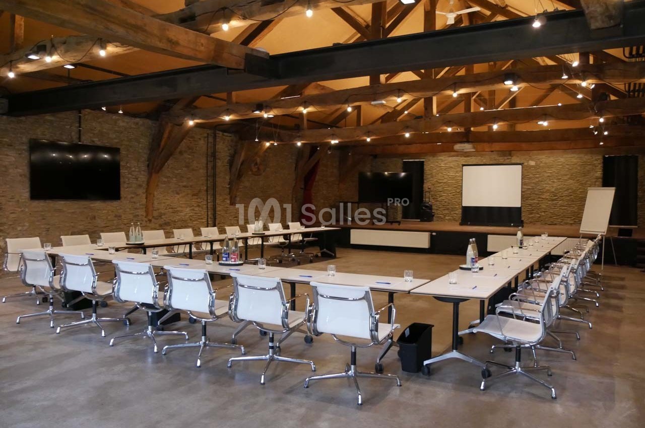 Salle de réunion spacieuse avec tables en U, chaises blanches, écran de projection et murs en pierre apparente.
