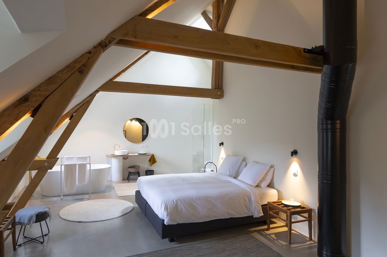 Chambre mansardée lumineuse avec poutres apparentes, lit double, baignoire, lavabo et poêle noir.
