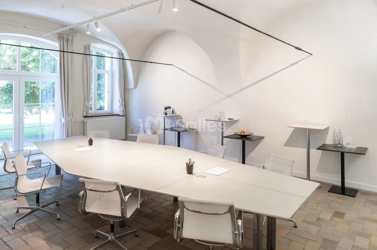 Salle de réunion lumineuse avec une grande table blanche, des chaises modernes et des fenêtres donnant sur un espace vert.
