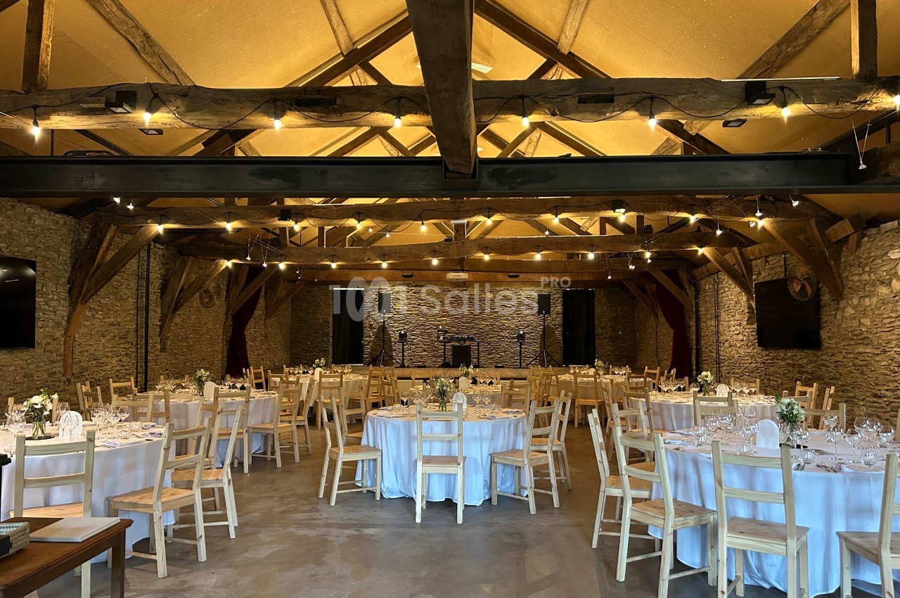 Salle de réception en pierre avec poutres apparentes, tables rondes dressées et chaises en bois disposées pour un événement.