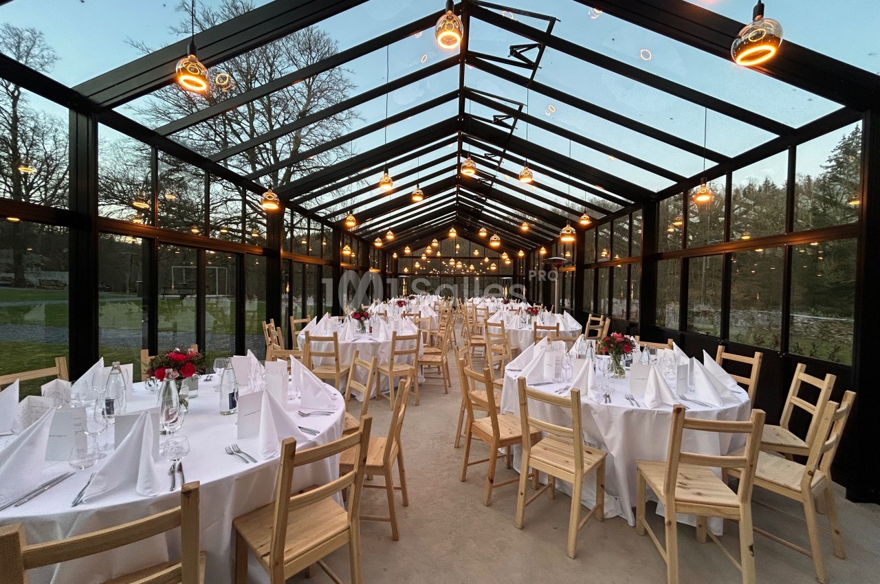 Salle de réception vitrée avec tables dressées, nappes blanches, chaises en bois et éclairage suspendu chaleureux.