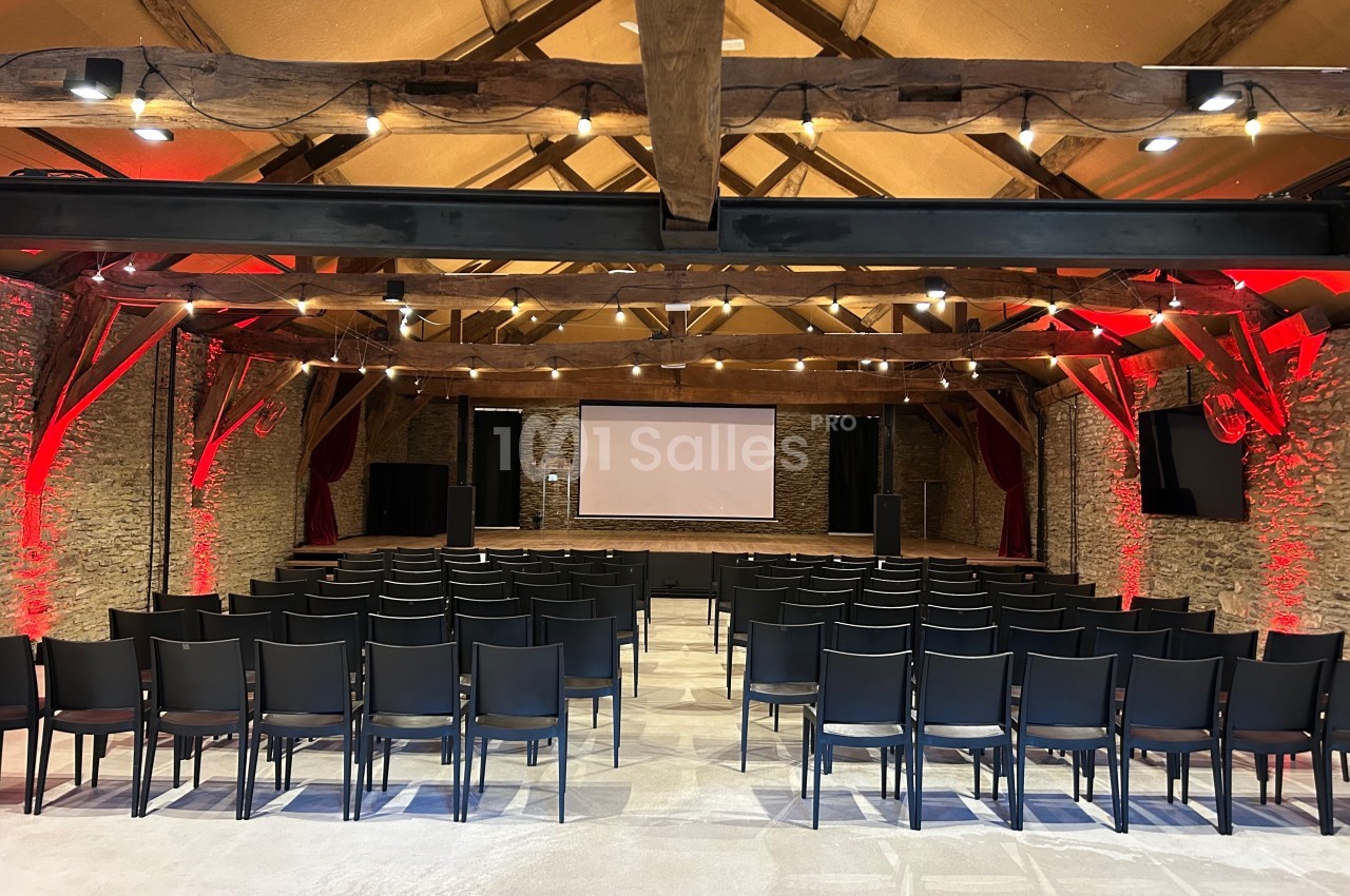 Salle de conférence avec des rangées de chaises noires, un écran de projection et des murs en pierre éclairés.