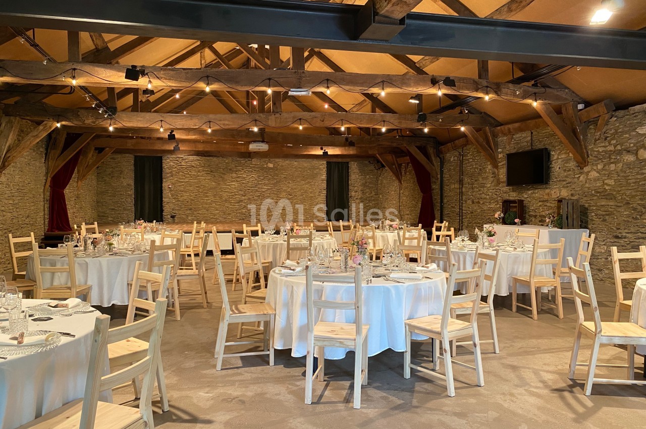 Salle de réception en pierre avec poutres apparentes, tables rondes dressées avec nappes blanches et chaises en bois clair.