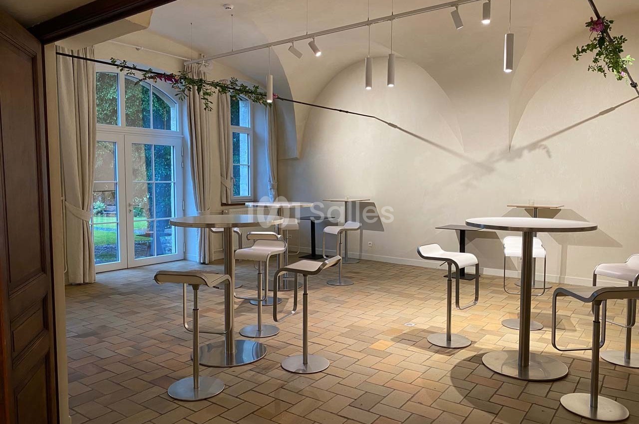 Salle lumineuse avec tables hautes, tabourets blancs et grandes fenêtres donnant sur un jardin verdoyant.