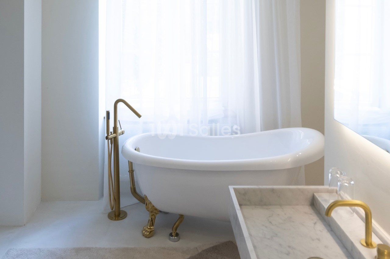 Salle de bain lumineuse avec baignoire sur pieds, robinetterie dorée et lavabo en marbre devant une grande fenêtre.