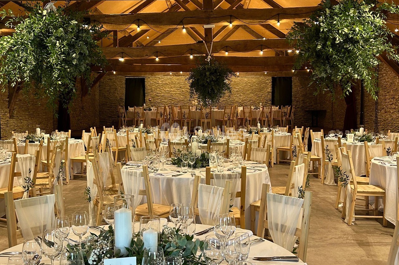Salle de réception décorée avec des tables rondes dressées, des chaises en bois et des suspensions végétales.
