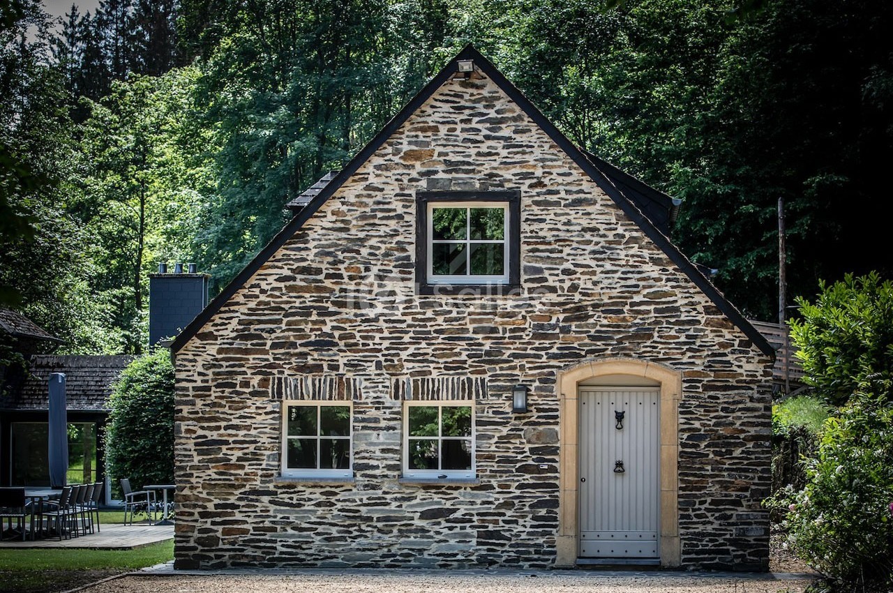 Façade d'une maison en pierre avec porte beige, fenêtres blanches et environnement boisé en arrière-plan.