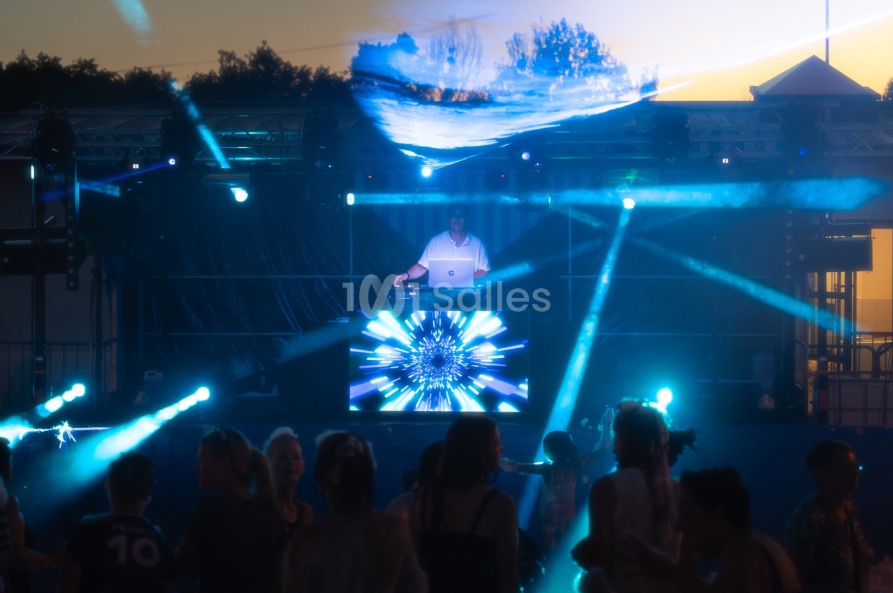 Scène de concert en plein air avec un DJ sur scène, des lumières colorées et une foule dansant au crépuscule.