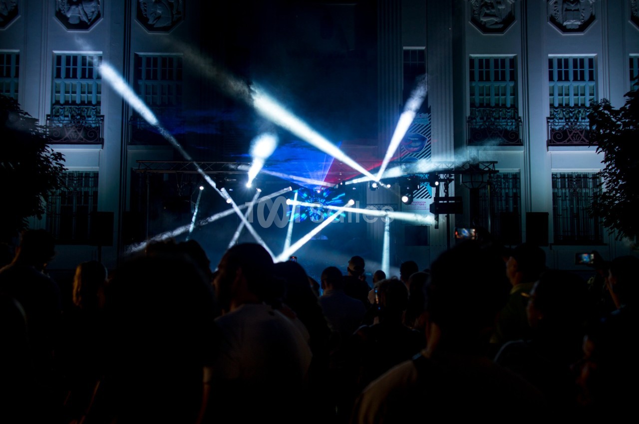 Foule devant un bâtiment illuminé, avec des faisceaux lumineux colorés traversant la scène lors d'un événement nocturne.
