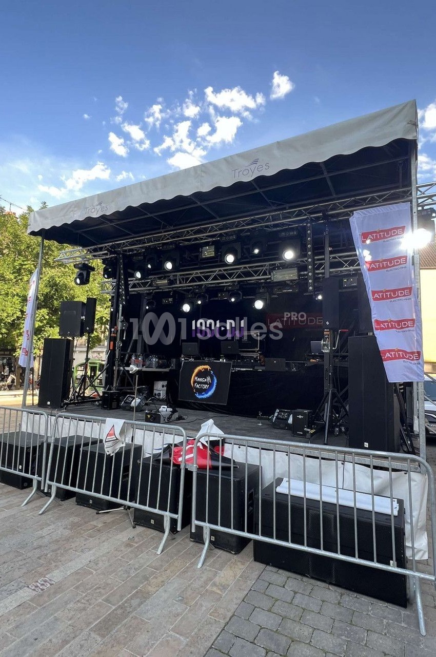 Scène extérieure équipée de matériel audio et éclairage, entourée de barrières, sur une place publique.