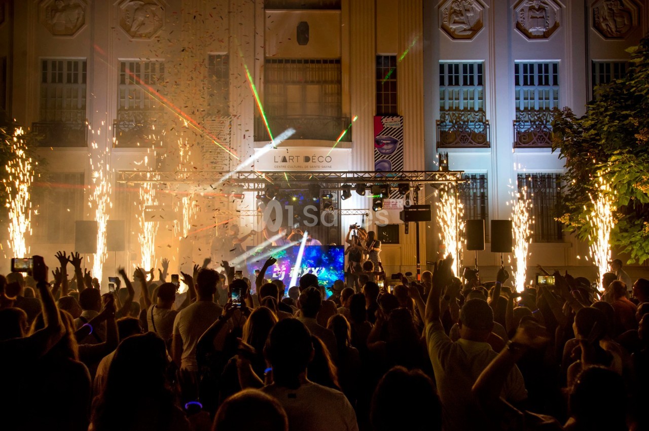 Foule en fête devant une scène illuminée par des feux d'artifice et des lasers, devant un bâtiment Art déco.
