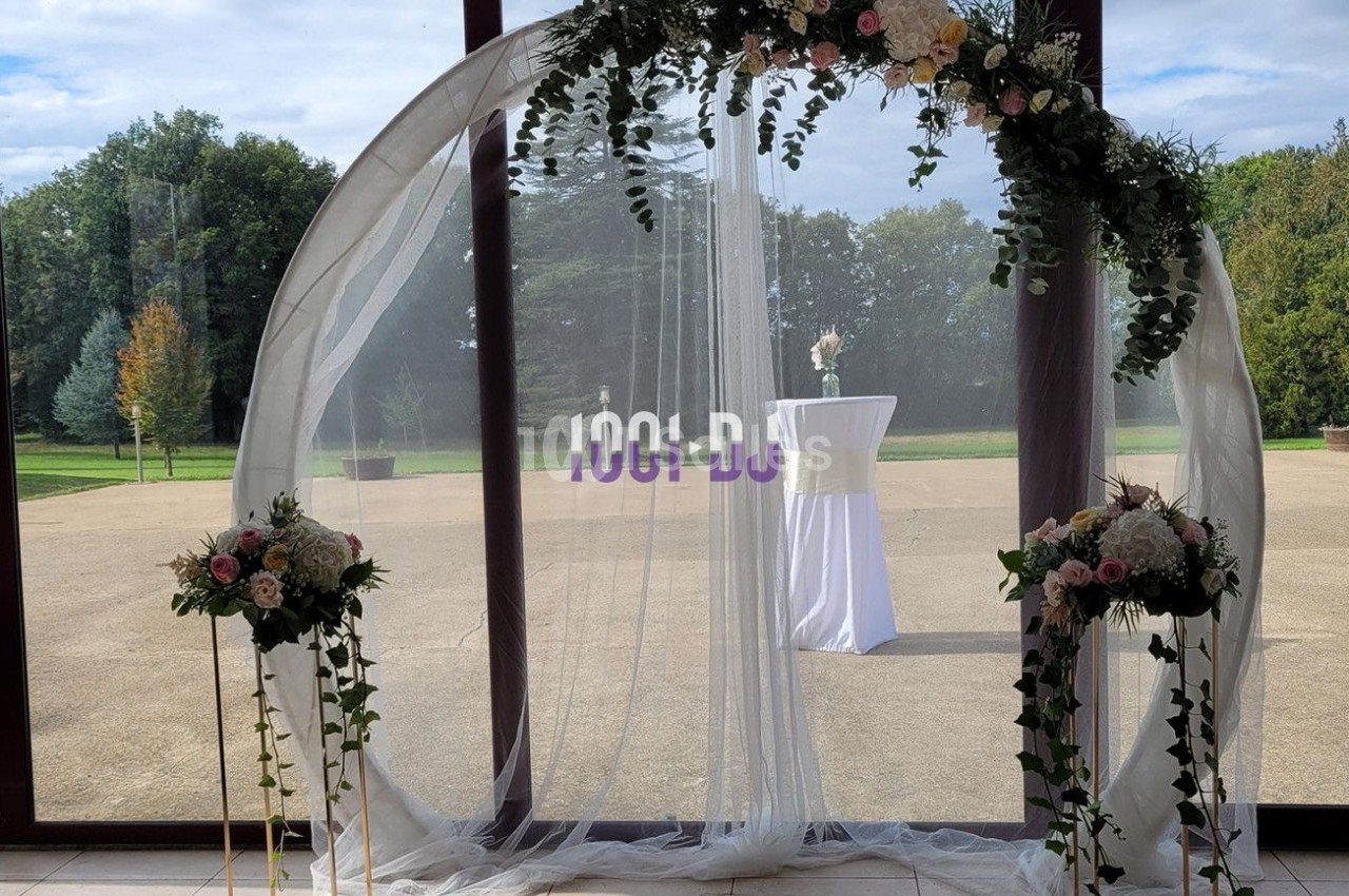 Arche de mariage circulaire décorée de fleurs et de voilages blancs, placée devant de grandes baies vitrées donnant sur un…