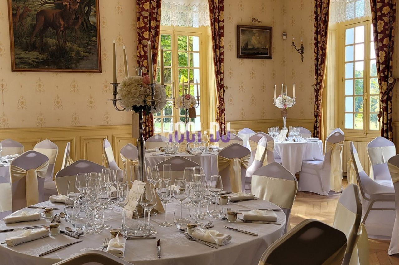 Salle de réception élégante avec tables rondes dressées, nappes blanches et décorations florales, éclairée par la lumière…
