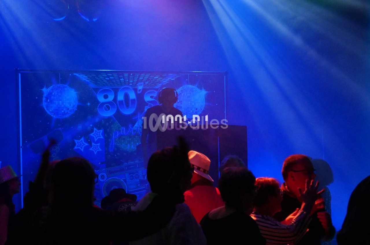 Personnes dansant dans une ambiance de fête rétro avec un DJ devant un décor sur le thème des années 80.