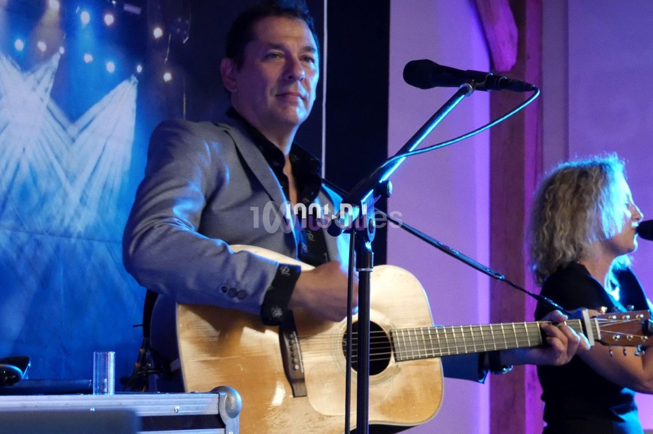 Un musicien joue de la guitare acoustique sur scène, accompagné d'une chanteuse au micro, sous des éclairages colorés.