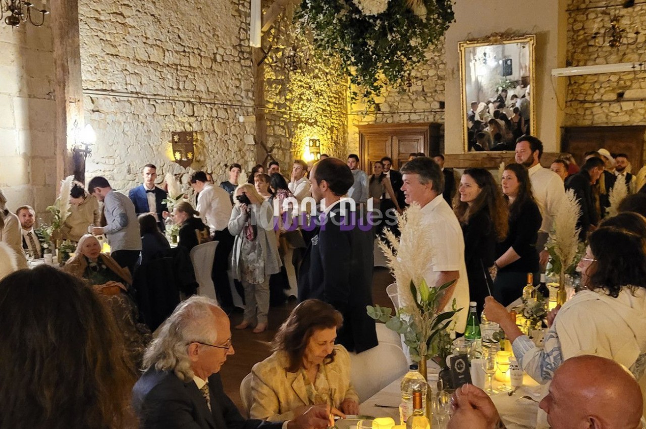 Salle de réception en pierre avec invités assis et debout, décorations florales suspendues et ambiance chaleureuse.