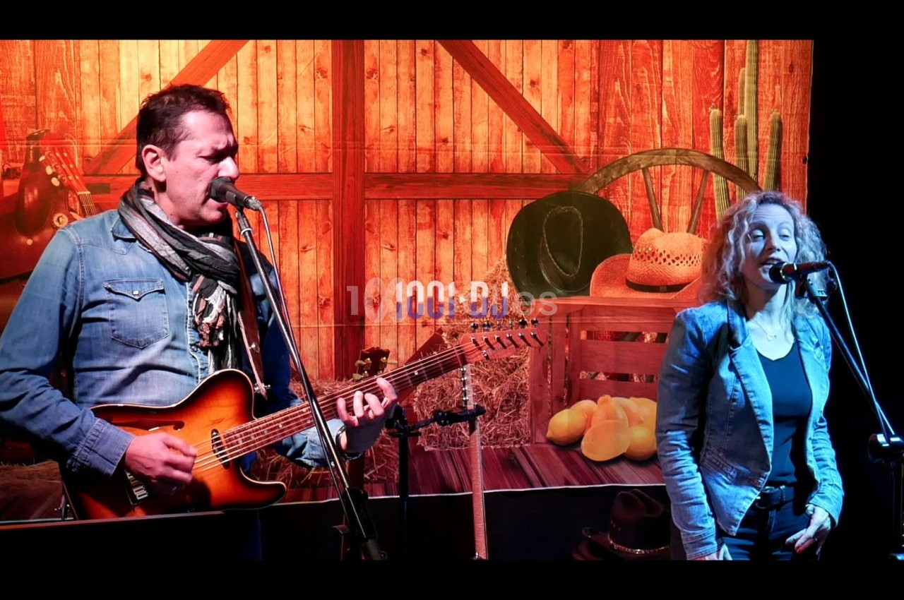 Un homme joue de la guitare et chante avec une femme sur une scène à thème western, décorée de bois et d'accessoires.