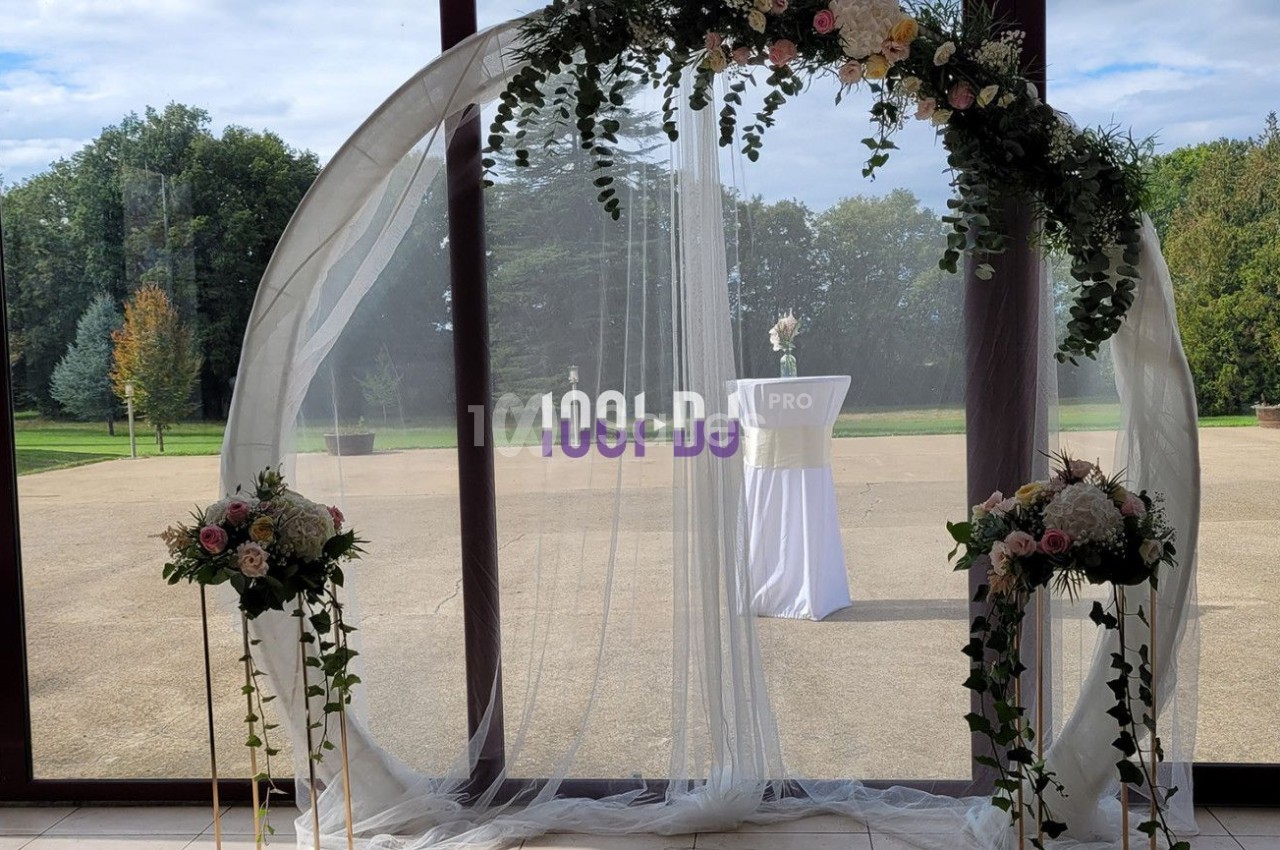 Arche de mariage circulaire décorée de fleurs et de voilages blancs, placée devant de grandes baies vitrées donnant sur un…
