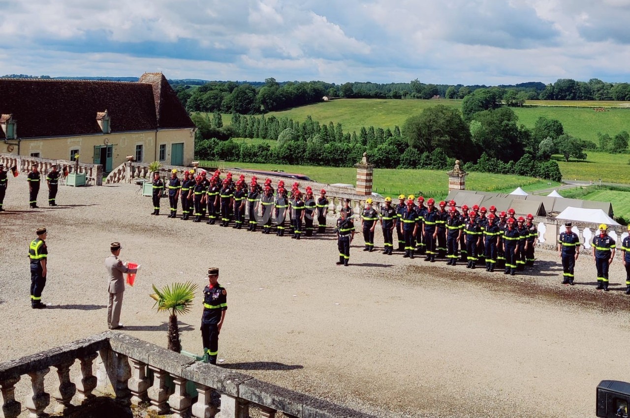 Des pompiers en uniforme alignés lors d'une cérémonie en extérieur, avec un bâtiment ancien et un paysage verdoyant en…