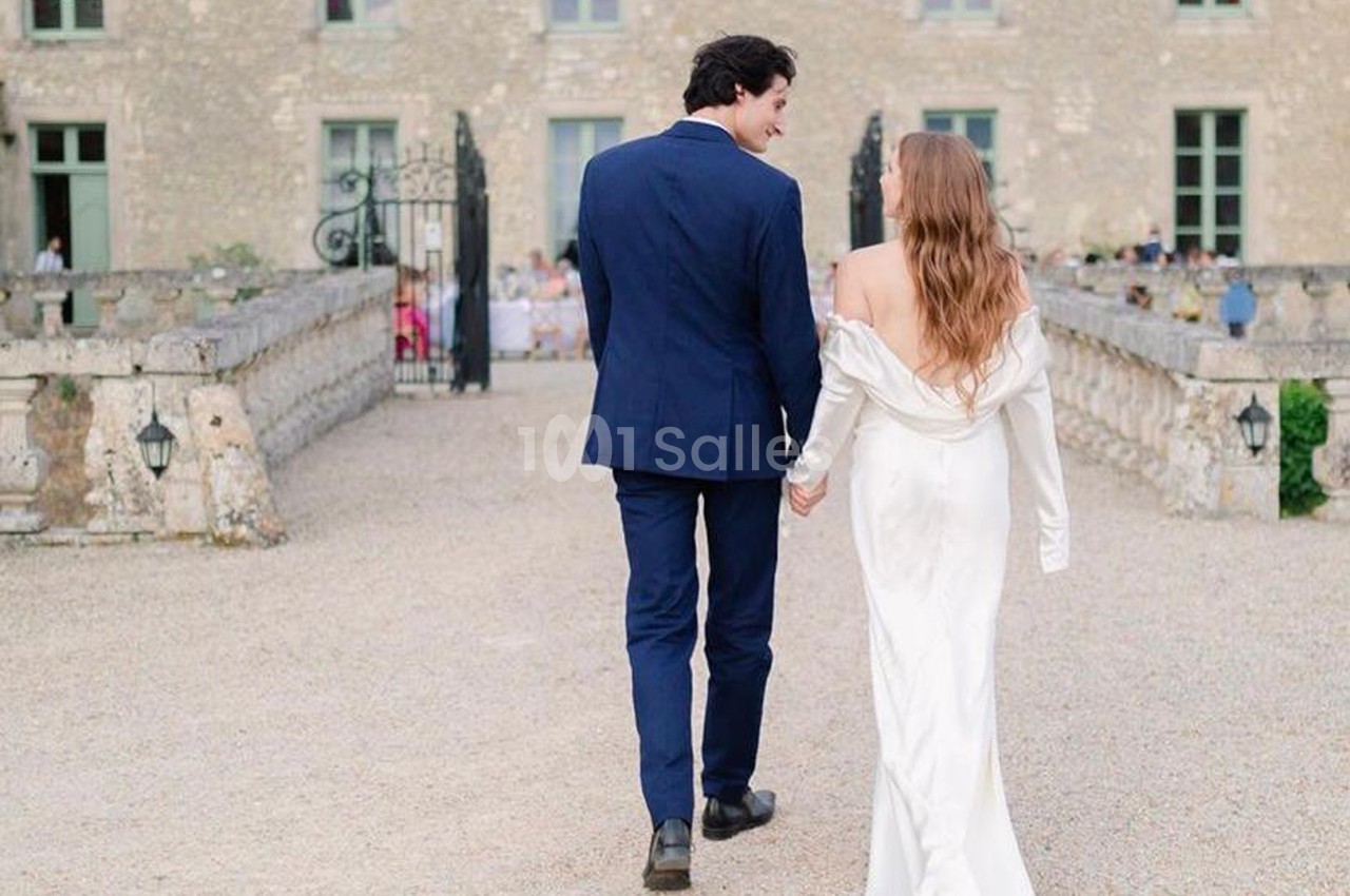 Un couple élégant marche main dans la main dans la cour d'un bâtiment en pierre avec des fenêtres et des portes vertes.
