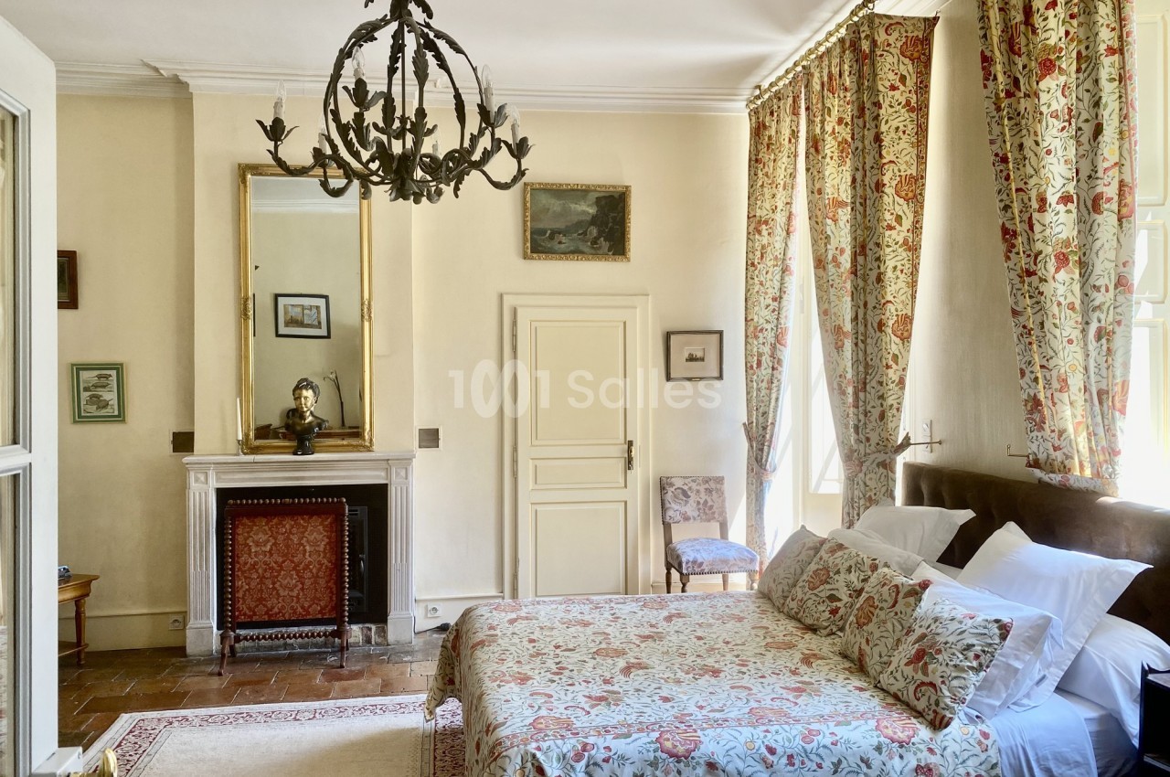 Chambre lumineuse avec lit double, cheminée, miroir doré, lustre en métal noir et rideaux à motifs floraux.