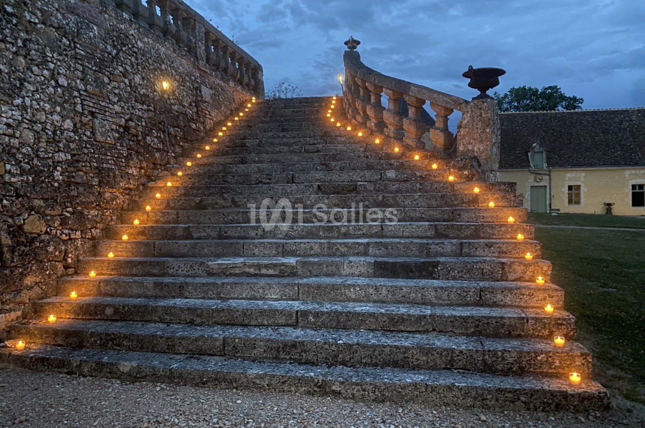 Escalier en pierre éclairé par des bougies alignées, menant à une balustrade, sous un ciel nuageux au crépuscule.
