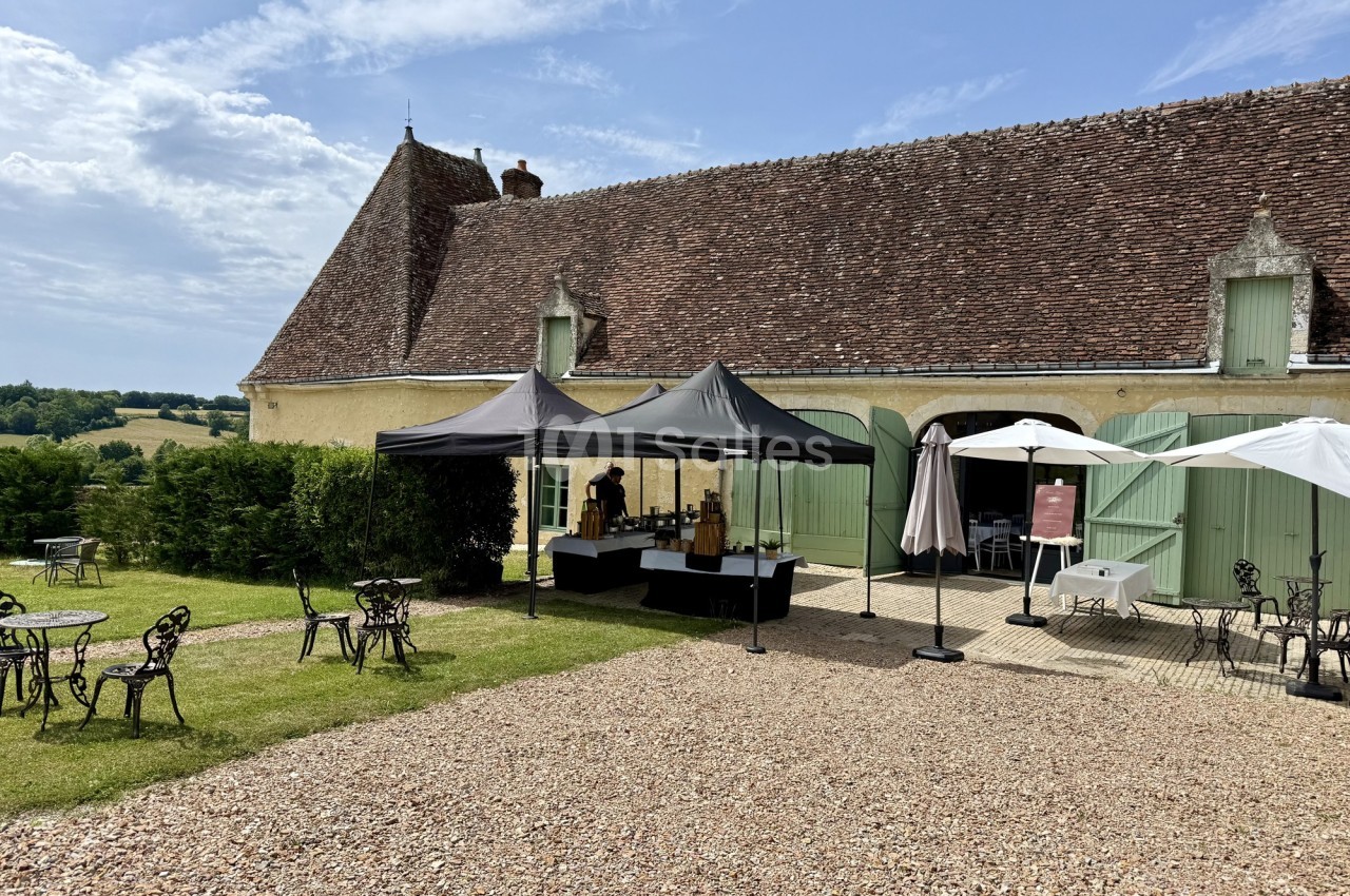 Cour extérieure avec une grande bâtisse en pierre, des tentes installées et des tables de jardin sur une pelouse.