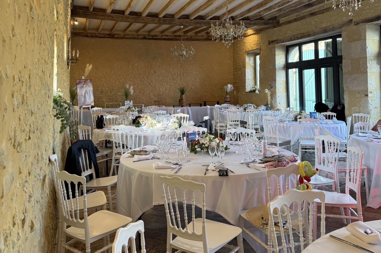Salle de réception décorée avec des tables rondes dressées, chaises blanches et murs en pierre apparente.