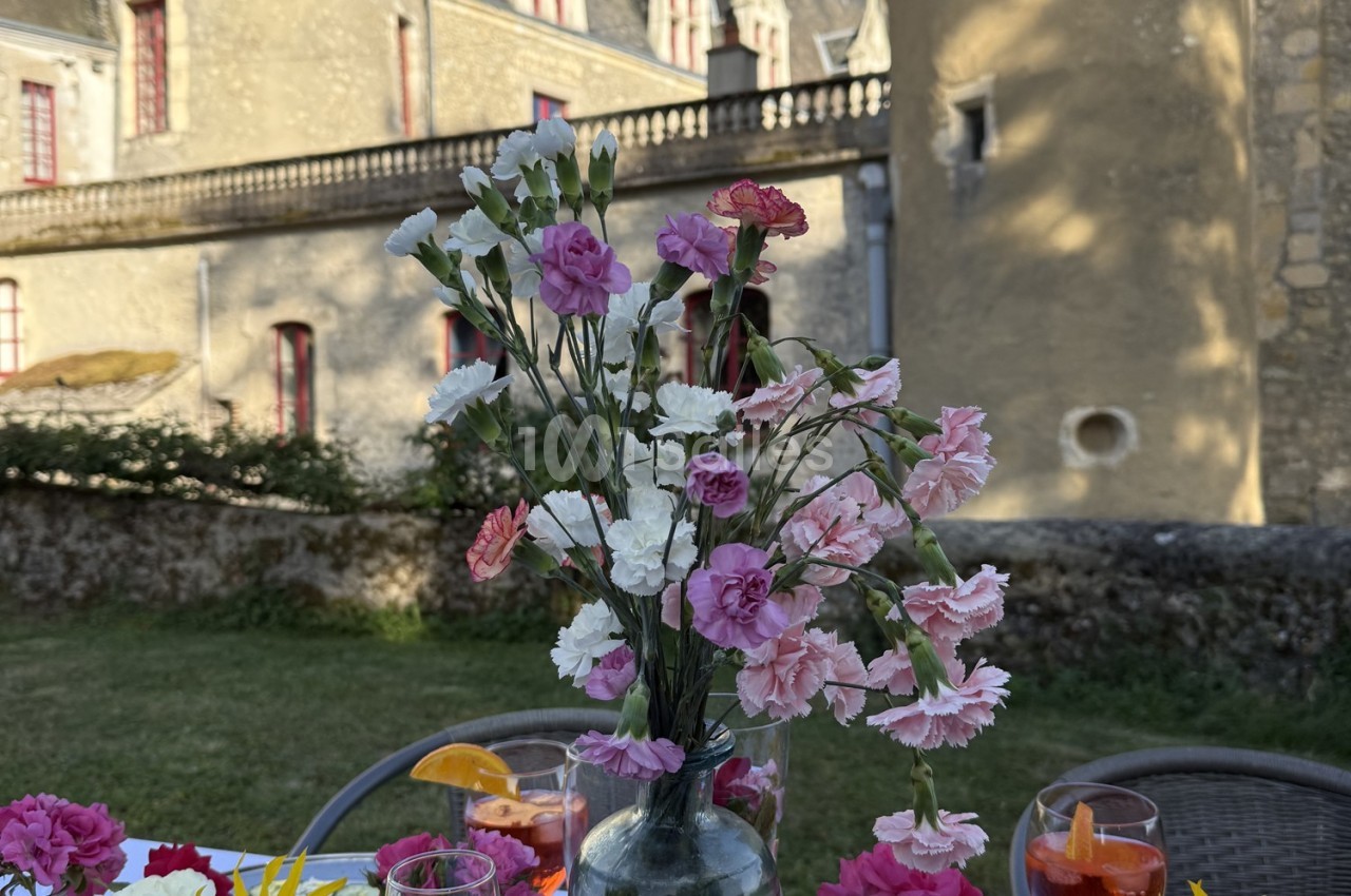 Vase de fleurs roses et blanches sur une table en extérieur, devant un bâtiment en pierre avec des fenêtres rouges.
