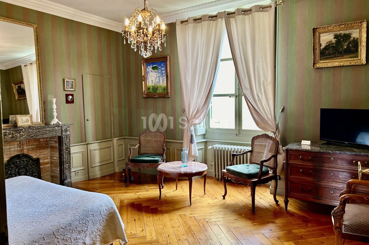 Chambre élégante avec parquet, cheminée, meubles anciens, lustre et grande fenêtre encadrée de rideaux blancs.