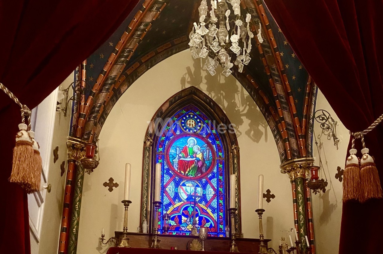 Vitrail coloré représentant une scène religieuse dans une chapelle ornée de rideaux rouges et d'un lustre.