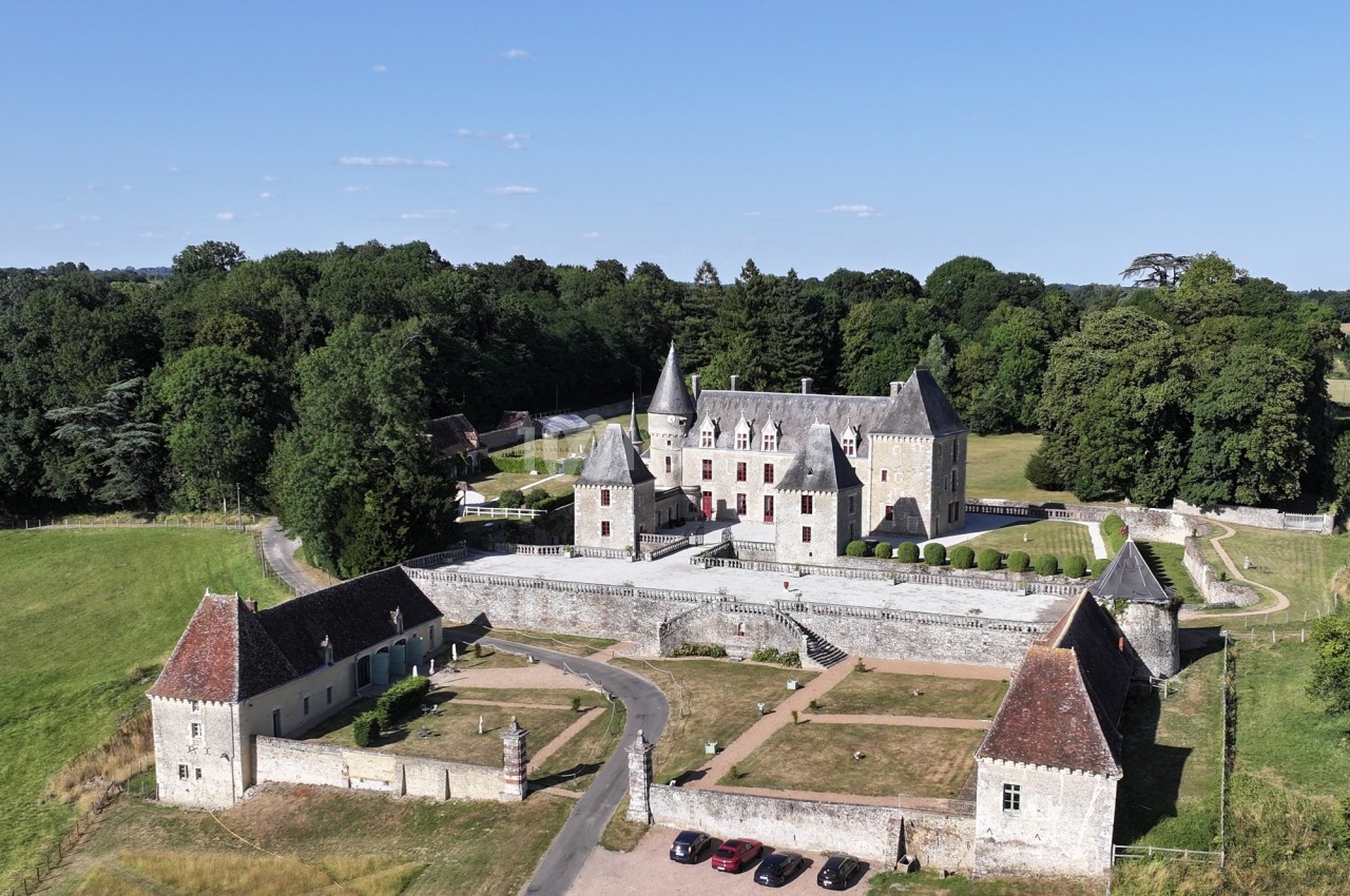 Vue aérienne d'un château entouré de verdure, avec des dépendances, une cour centrale et des voitures stationnées.