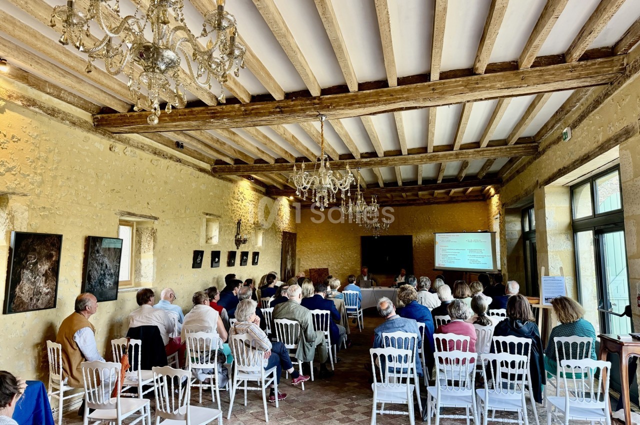 Des personnes assistent à une présentation dans une salle avec murs en pierre, poutres apparentes et chaises blanches.