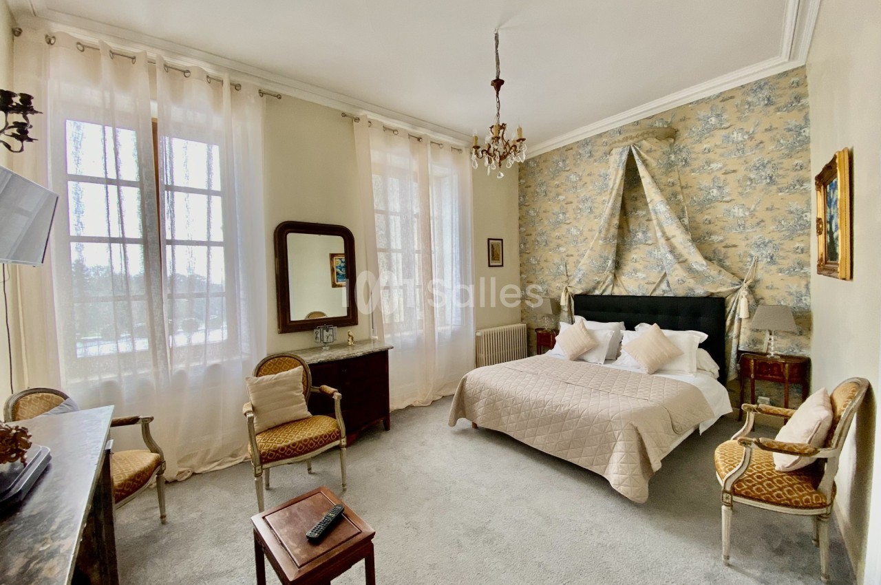 Chambre lumineuse avec lit double, mobilier ancien, murs clairs, rideaux transparents et vue sur l'extérieur.