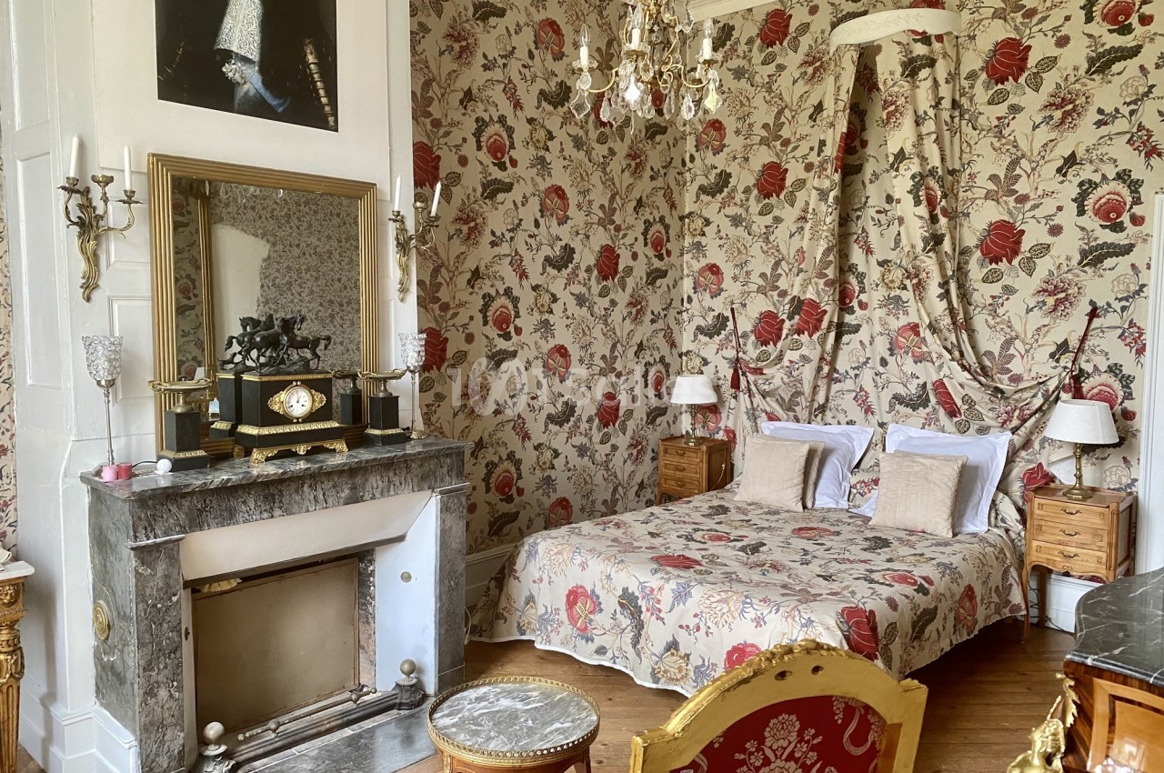 Chambre décorée de motifs floraux avec lit à baldaquin, cheminée en marbre, mobilier ancien et lustre doré.