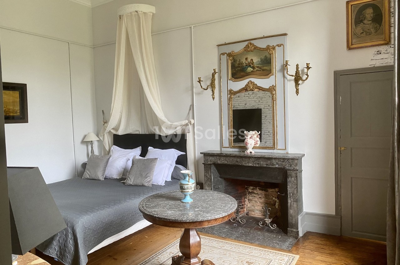 Chambre élégante avec lit à baldaquin, cheminée en marbre, miroir doré et table ronde sur un tapis.