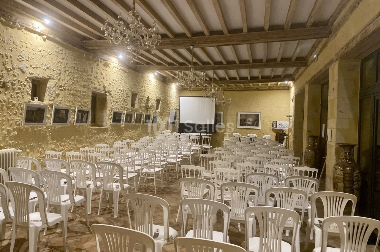 Salle en pierre avec des rangées de chaises blanches disposées face à un écran de projection.