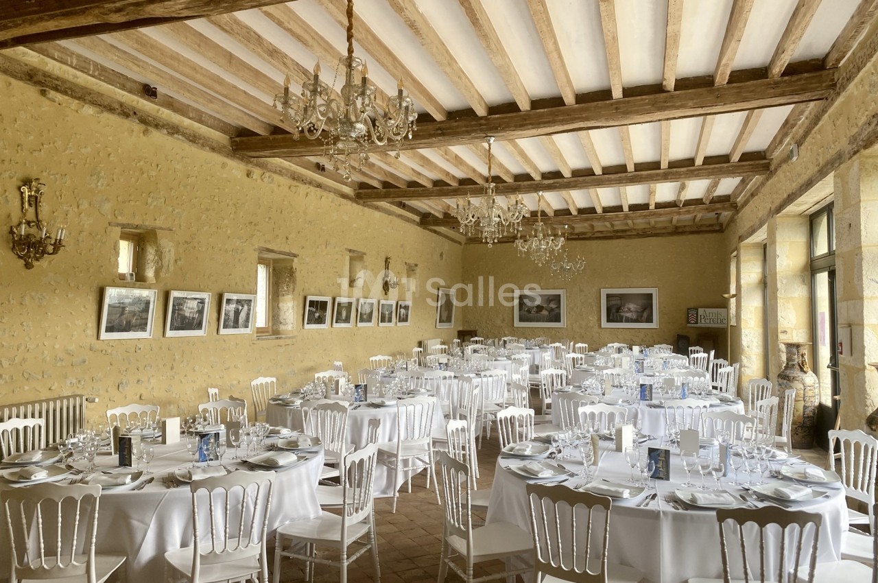 Salle de réception lumineuse avec tables rondes dressées, chaises blanches et murs en pierre ornés de cadres.