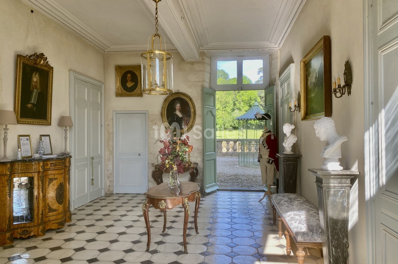 Intérieur d'une entrée lumineuse avec sol en damier, meubles anciens, portraits et vue sur un jardin.