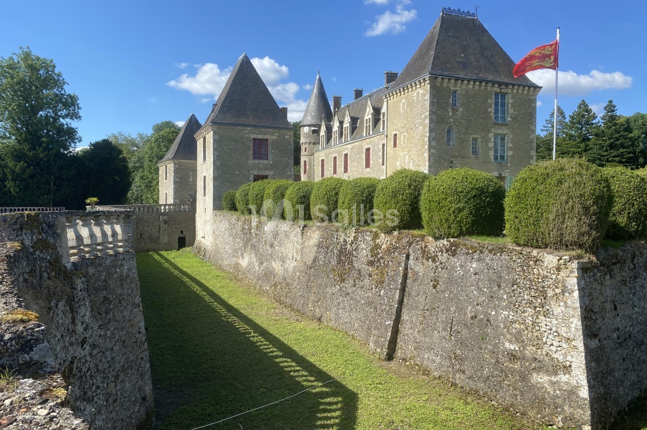 Château en pierre entouré de douves sèches, avec drapeau rouge et jaune flottant sur une tour, sous un ciel bleu.