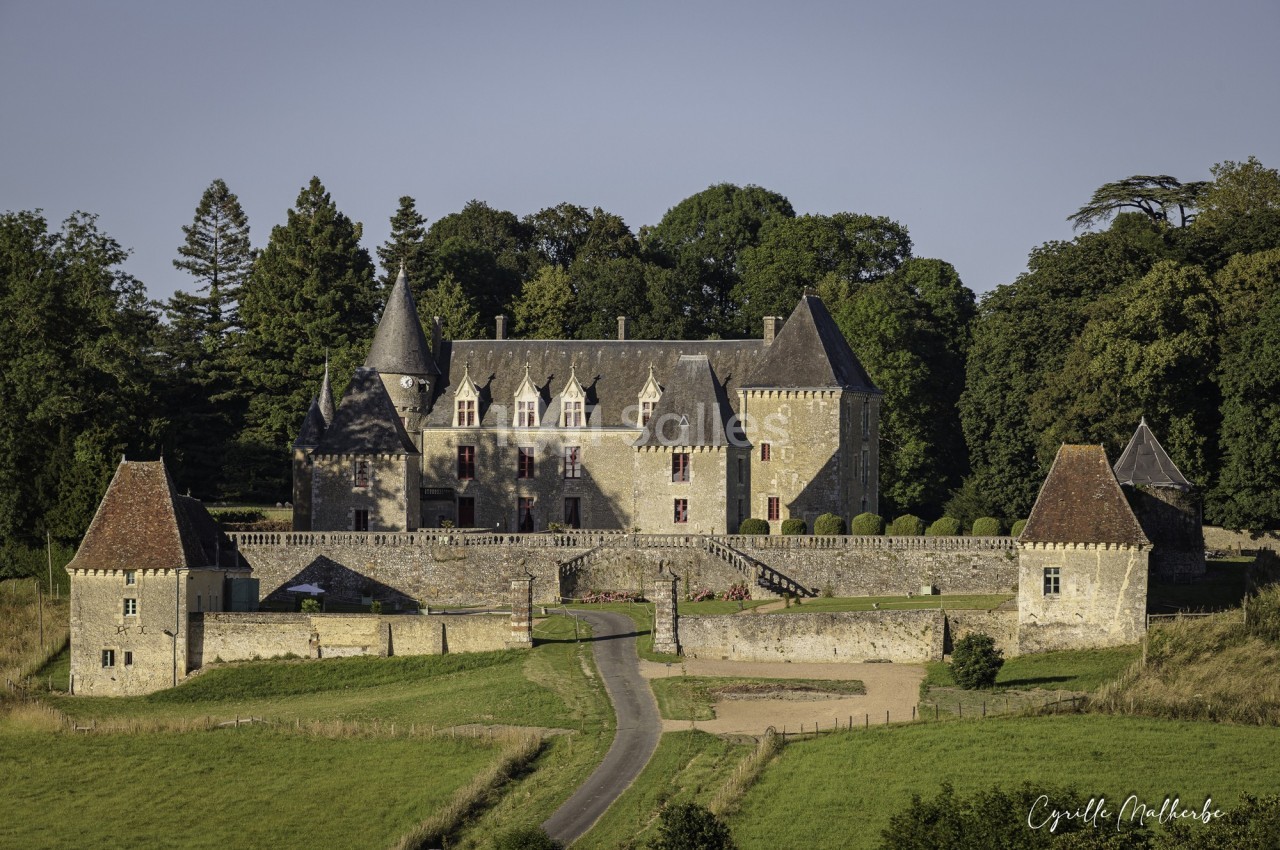 Château en pierre entouré de verdure, avec tours et toits en ardoise, vu sous un ciel dégagé.