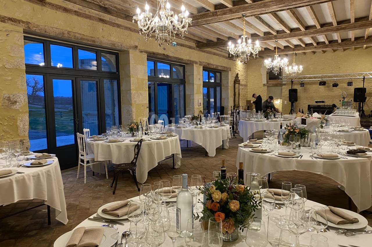 Salle de réception élégante avec tables dressées, chandeliers suspendus et grandes fenêtres donnant sur l'extérieur.