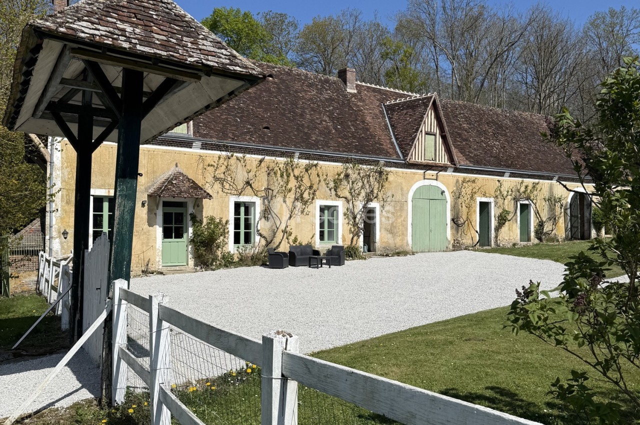 Façade d'une maison ancienne en pierre avec toit en tuiles, entourée de verdure et d'une cour gravillonnée.